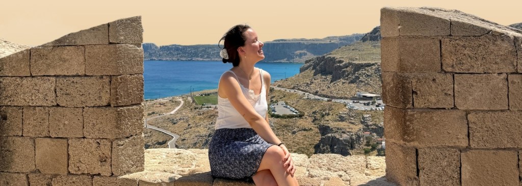 Die Autorin sitzt entspannt auf einer Steinmauer mit Blick auf eine Küstenlandschaft, während sie die Sonne genießt.