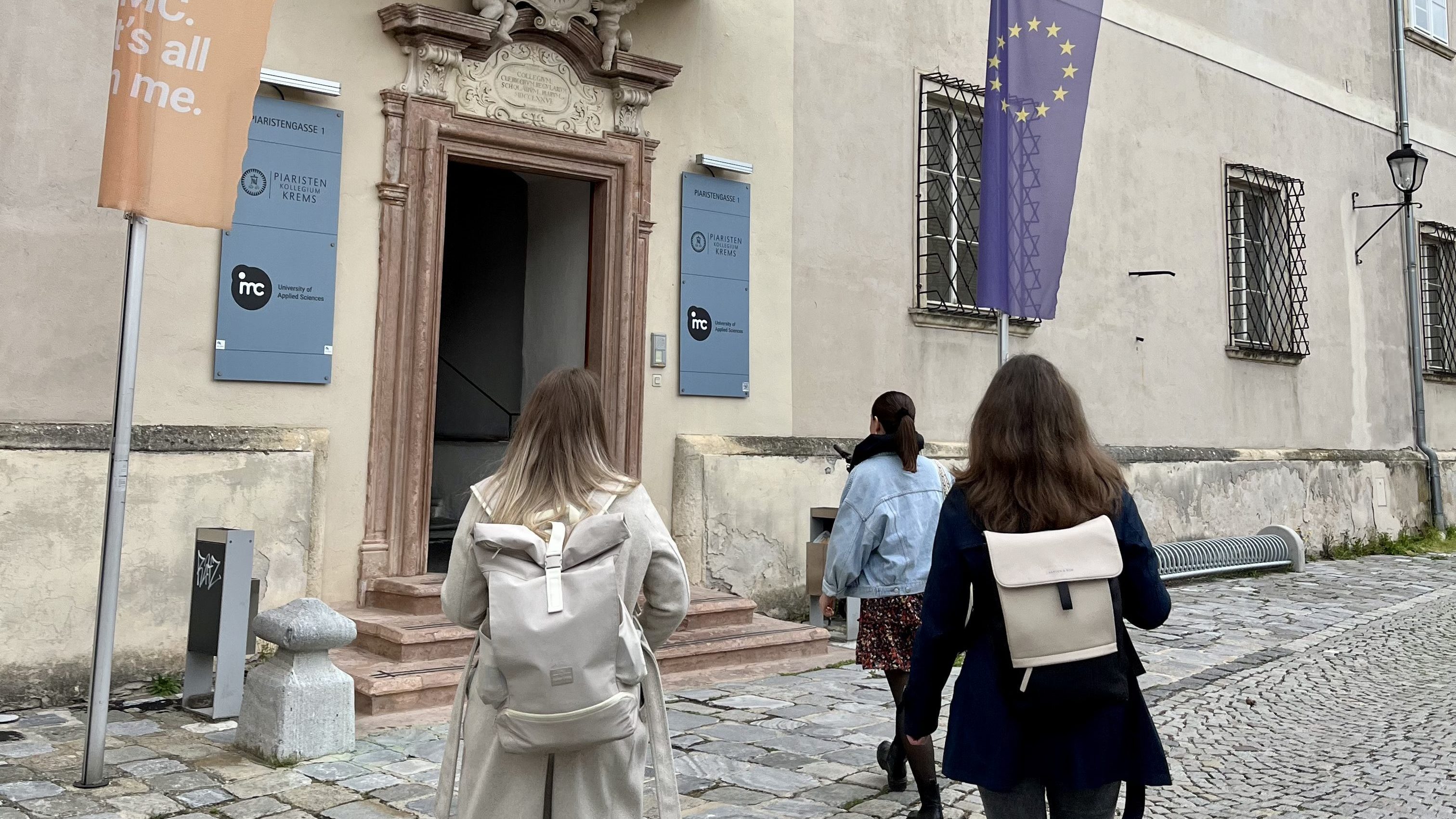 Die Autorin geht mit zwei Freundinnen mit Rucksäcken auf einem gepflasterten Gehweg zur Eingangstür eines Gebäudes mit einem Schild der Universität für Angewandte Wissenschaften.