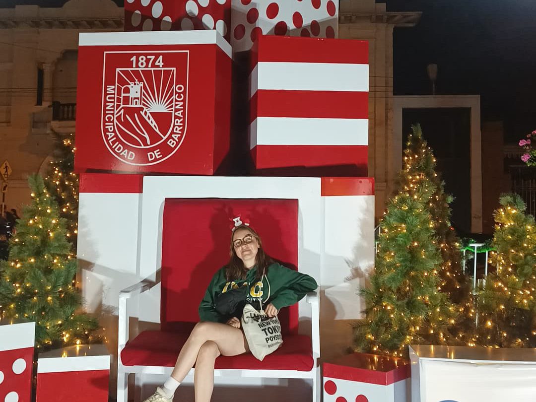 Eine Person sitzt in einem großen, roten und weißen Weihnachtsstuhl vor einer festlich dekorierten Kulisse mit Weihnachtsbäumen, Geschenkspäckchen und Lichtern im Viertel Barranco in Lima, Peru.