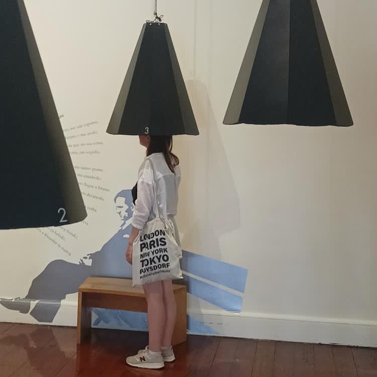 Eine Person steht in einem Raum mit grauen Pendelleuchten über dem Kopf. Im Hintergrund sind Schriftzüge und eine künstlerische Darstellung an der Wand zu sehen.