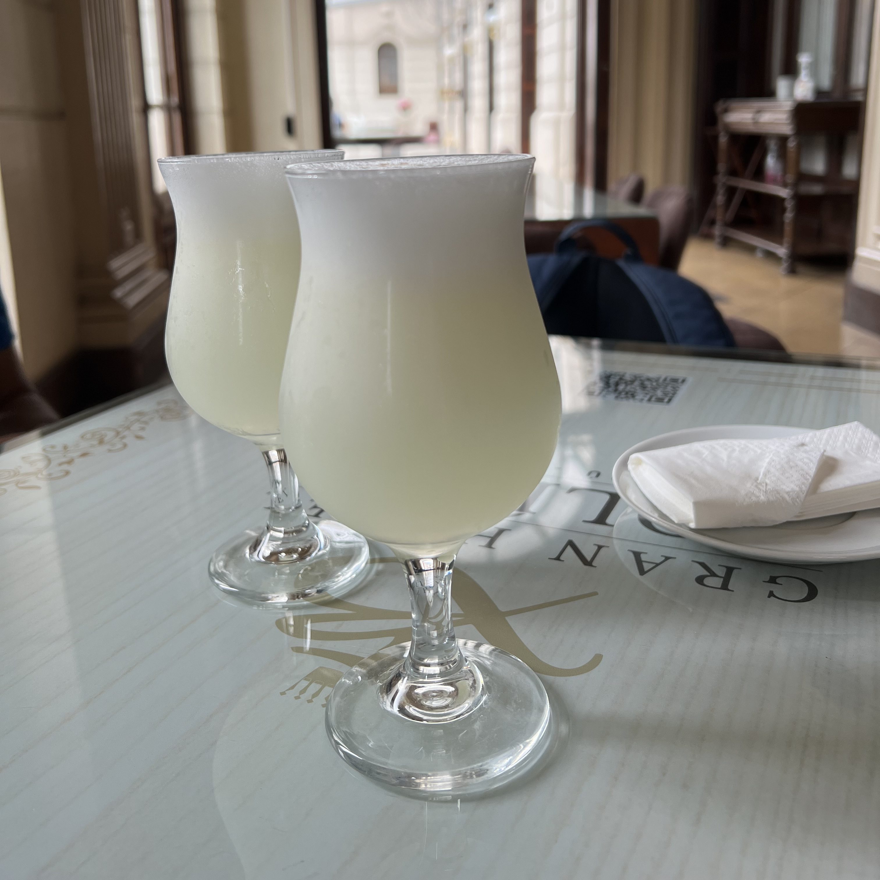 Zwei Gläser Pisco Sour auf einem Tisch in einem stilvollen Restaurant. Die Gläser sind elegant und enthalten einen schaumigen Cocktail, während im Hintergrund ein heller Raum mit Fenstern zu sehen ist.