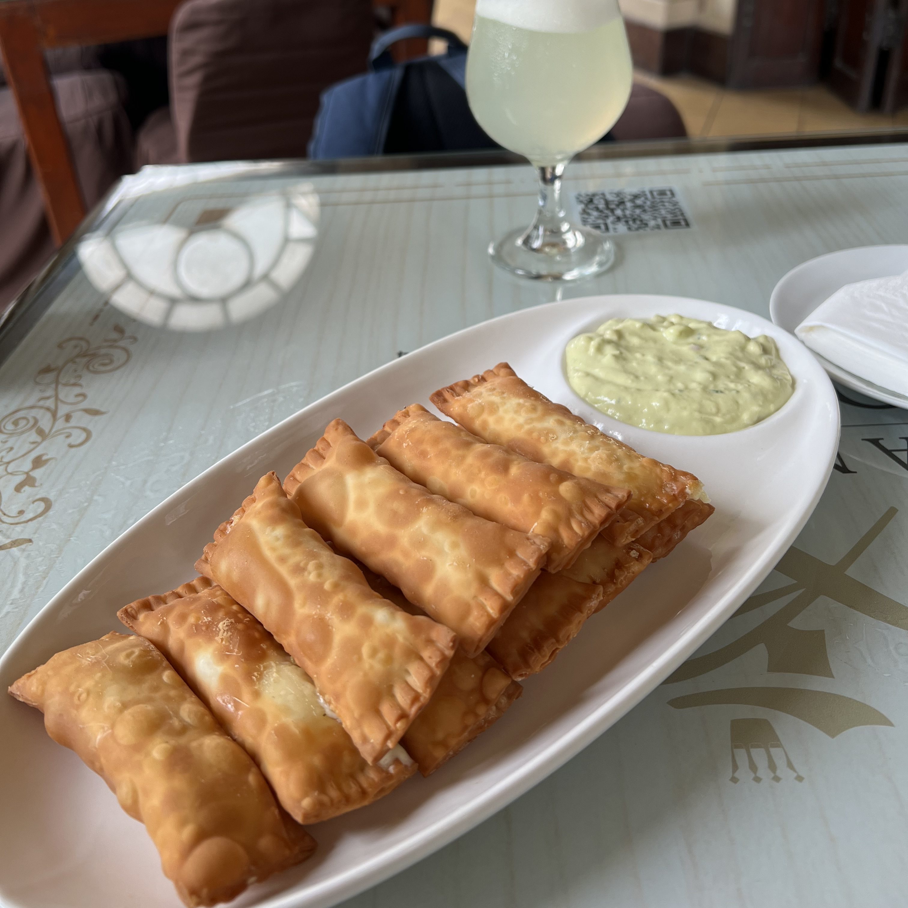 Ein Teller mit frittierten Tequeños, serviert mit einer cremigen Avocadosauce, und einem Glas Pisco Sour im Hintergrund, auf einem Tisch.