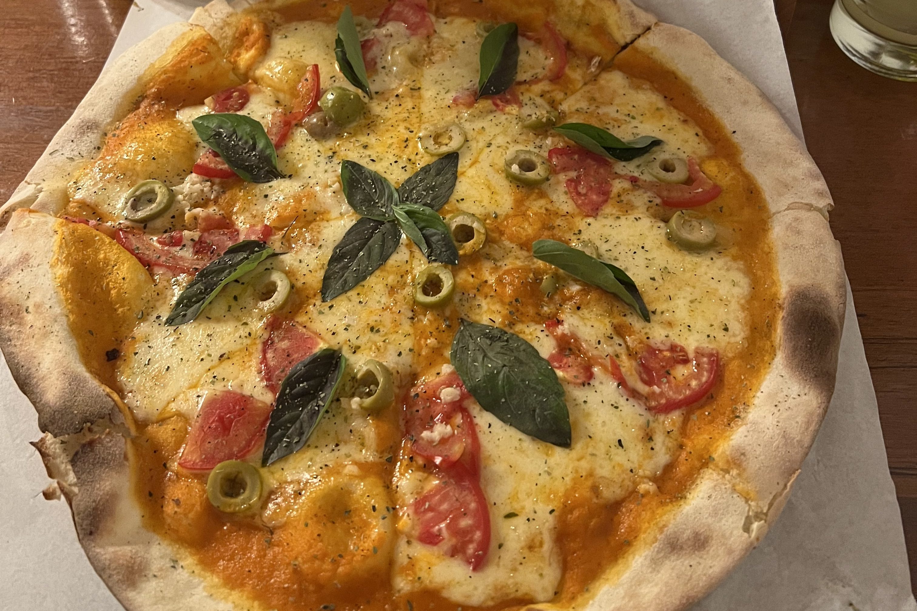 Eine frisch gebackene Pizza belegt mit Tomaten, Oliven und Basilikum auf einem Holzbrett serviert.